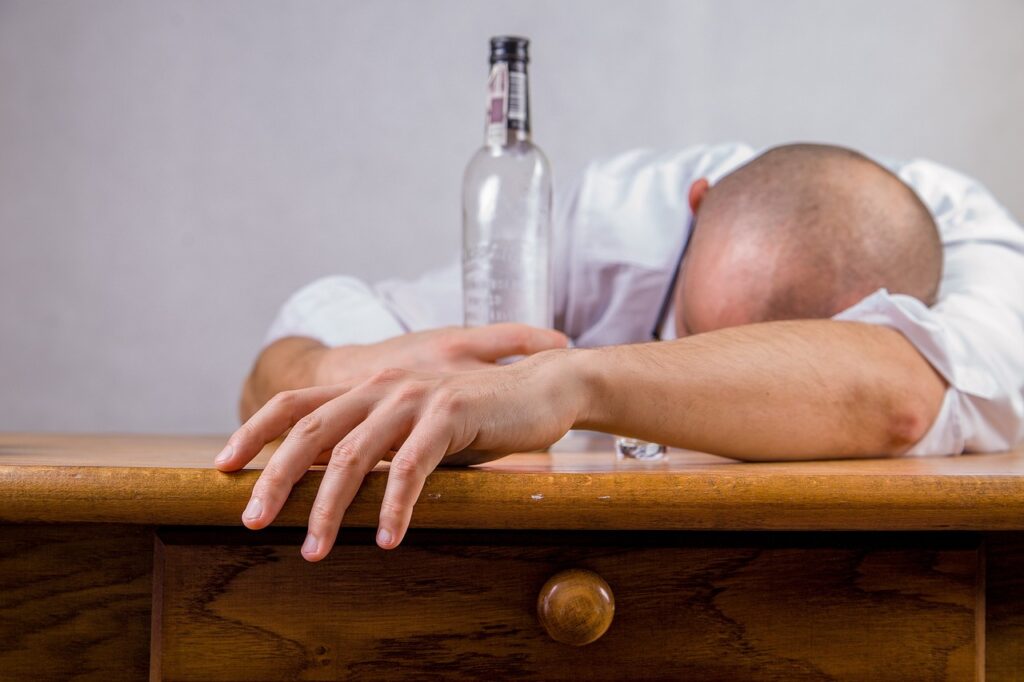 Man lying face down over a desk, appearing asleep with a bottle of liquor in his hand.